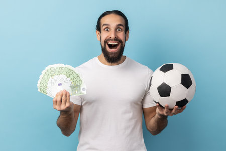 Portrait of man with beard wearing white T-shirt holding soccer ball and hundred euro bills, screaming looking camera, betting and winning. Indoor studio shot isolated on blue background.の写真素材