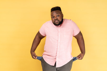 Portrait of frustrated worried man wearing pink shirt turning out empty pockets showing no money gesture, frowning face. Indoor studio shot isolated on yellow background.の写真素材