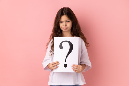 Portrait of winsome beautiful little girl wearing white T-shirt looking at camera, holding paper with question mark, thinks about tasks. Indoor studio shot isolated on pink background.の写真素材