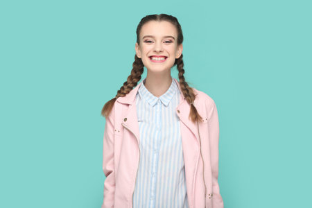 Portrait of extremely happy joyful teenager girl with braids wearing pink jacket standing looking at camera with toothy smile, smiles broadly. Indoor studio shot isolated on green background.の写真素材