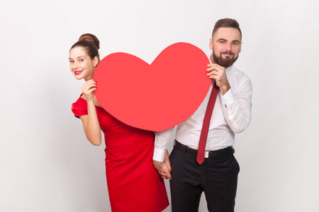Portrait of smiling satisfied man in white shirt and woman in red dress standing together, peeping out from big heart, expressing love. Indoor studio shot isolated on gray background.の写真素材