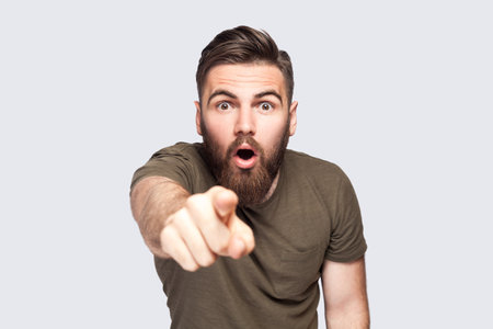 Surprised wondered bearded man wearing dark green T-shirt pointing fingers looking at camera with open mouth and big eyes, shocked with choice. Indoor studio shot isolated on light gray background.の写真素材