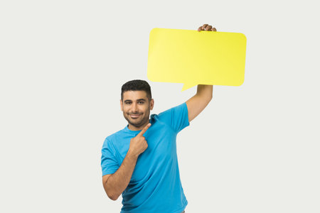 Portrait of optimistic smiling unshaven man wearing blue T- shirt standing pointing at yellow speech...
