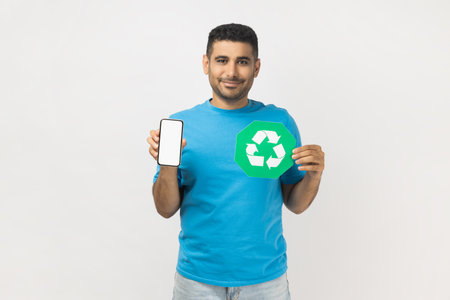 Portrait of happy unshaven man wearing blue T- shirt standing standing holding recycling sign and smart phone with blank screen for advertisement. Indoor studio shot isolated on gray background.の写真素材