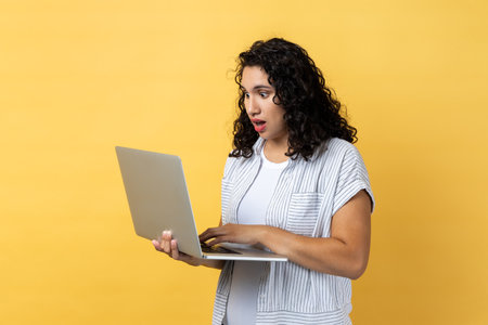 Portrait of shocked astonished woman with dark wavy hair working on laptop, looking at screen with big eyes and open mouth. Indoor studio shot isolated on yellow background.の写真素材