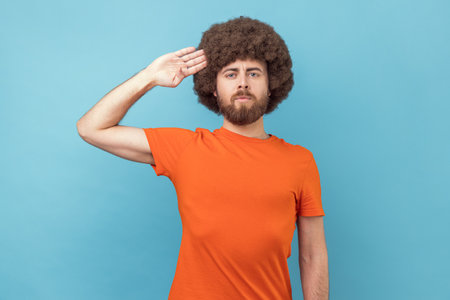 Portrait of man with Afro hairstyle wearing orange T-shirt saluting with respect as if soldier...