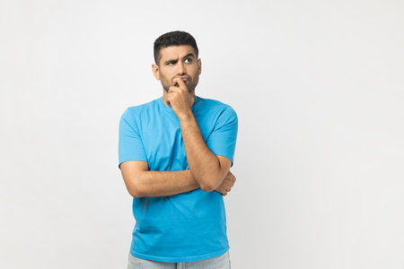 Portrait of serious thoughtful unshaven man wearing blue T- shirt standing keeps hand on his chin, thinking about his work, looking away. Indoor studio shot isolated on gray background.の写真素材