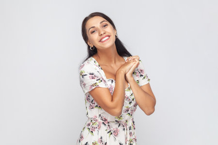 Portrait of cute charming beautiful woman wearing white dress looking at camera with toothy smile, keeps hands together, admires. Indoor studio shot isolated on gray background.の写真素材