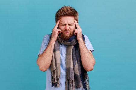 Portrait of bearded man wrapped in scarf standing with painful grimace, touching sore head, suffering migraine, sick with influenza. Indoor studio shot isolated on blue background.の写真素材