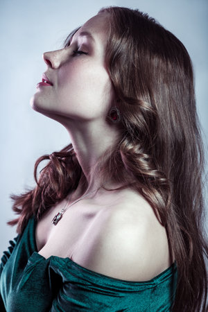 Side view portrait of attractive adorable woman with long brown hair wearing green dress, standing with closed eyes, has calm relaxed look. Indoor studio shot isolated on gray background.の写真素材