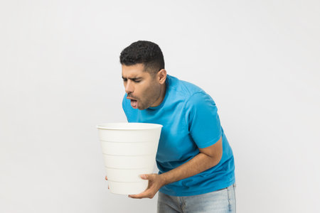 Portrait of sick unhealthy ill unshaven man wearing blue T- shirt standing suffering stomachache, feels nausea and vomits, holding bin in hands. Indoor studio shot isolated on gray background.の写真素材