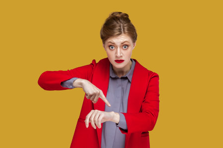 You are late. Portrait of serious strict woman with red lips standing pointing finger to her smartwatch, looking at camera, wearing red jacket. Indoor studio shot isolated on yellow background.の写真素材