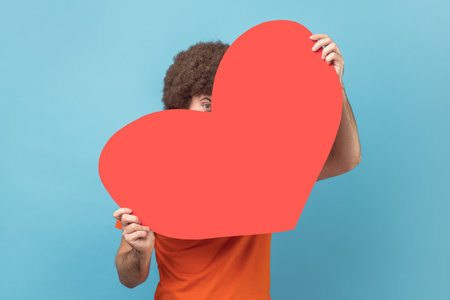 Portrait of man hiding face behind big red heart and looking at camera with curious prying eyes, expressing i love you, feeling affection fondness. Indoor studio shot isolated on blue background.の写真素材