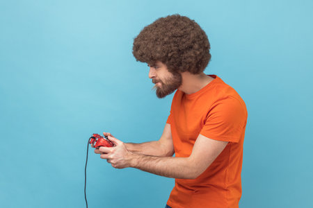 Side view of concentrated man with Afro hairstyle wearing orange T-shirt holding in hands red gamepad joystick, grimacing playing video games. Indoor studio shot isolated on blue background.の写真素材