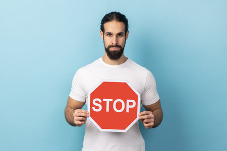 Portrait of serious man with beard wearing white T-shirt holding red Stop sign, looking at camera with negative aggressive expression, showing ban. Indoor studio shot isolated on blue background.の写真素材