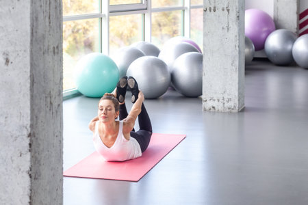 Portrait of sporty and flexible woman lying on bellies and holding legs in hands while practicing yoga on exercise mat, keeps eyes closed. Indoor shot with window on background.の写真素材