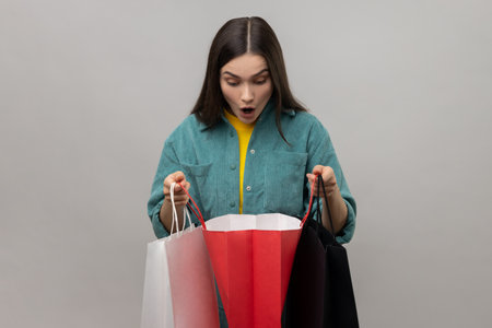 Excited amazed woman with dark hair looking inside shopping bags, having shocked expression, being surprised, wearing casual style jacket. Indoor studio shot isolated on gray background.の写真素材
