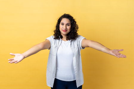 Please, take for free. Portrait of generous friendly woman with dark wavy hair welcoming with wide open arms and smiling kindly, happy to embrace you. Indoor studio shot isolated on yellow background.の写真素材
