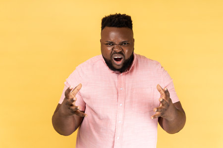 Portrait of angry aggressive man wearing pink shirt standing with raised arms, expressing aggression, screaming, frowning face. Indoor studio shot isolated on yellow background.の写真素材