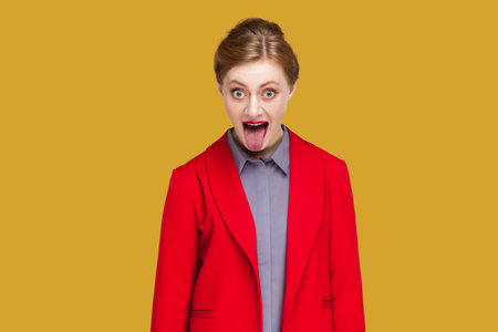 Portrait of woman with red lips sticks out tongue, makes happy grimace, has long hair, expresses positive emotions, wearing red jacket. Indoor studio shot isolated on yellow background.の写真素材
