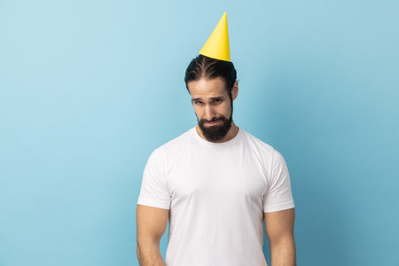 Portrait of sad unhappy man with beard wearing white T-shirt and party cone standing alone, nobody came to his birthday party, expressing sadness. Indoor studio shot isolated on blue background.の写真素材