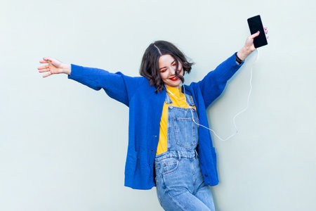 Portrait of joyful hipster woman in denim overalls yellow T-shirt and blue jacket listening to music on smart phone, keeps eyes closed, enjoying. Indoor studio shot isolated on light green backgroundの写真素材