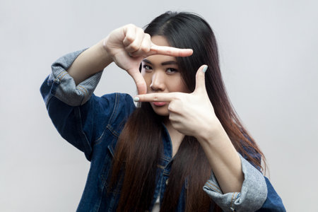 Portrait of serious concentrated attractive brunette woman in blue denim jacket standing looking at camera and gesturing finger frame. Indoor studio shot isolated on gray background.の写真素材