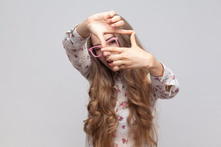 Woman in glasses with wavy blond hair gesturing picture frame with hands, looking through fingers and focusing on interesting moment, taking photo. Indoor studio shot isolated on gray background.の写真素材