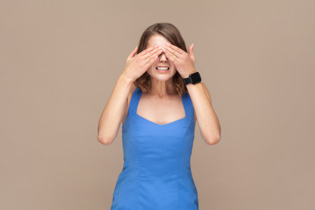 Dont want to look at this. Portrait of scared woman with wavy hair standing, covering eyes with hand, refusing to watch, wearing blue dress. Indoor studio shot isolated on light brown background.の写真素材