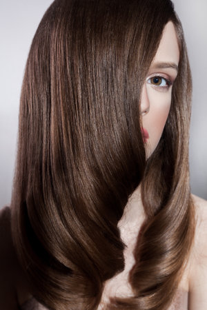 Portrait of attractive brunette woman fashion model with silk straight shiny hair and makeup, covering half of face with hair, looking at camera. Indoor studio shot isolated on gray background.の写真素材
