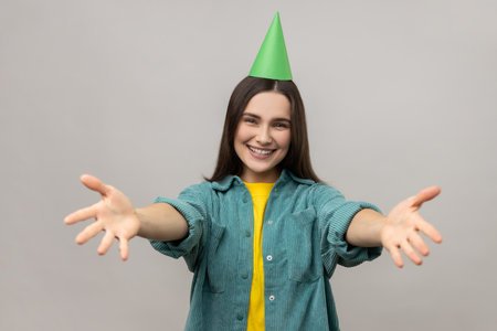 Woman with party cone keeping hands wide open to embrace, giving warm welcome, greeting and sharing love, wearing casual style jacket. Indoor studio shot isolated on gray background.の写真素材