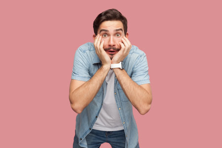 Portrait of shocked astonished bearded man in blue casual style shirt standing with hands on cheeks,...