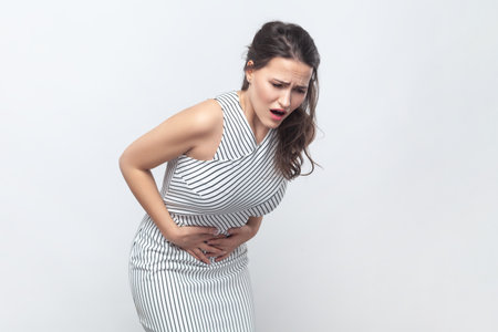 Portrait of sick unhealthy ill brunette woman touching her painful belly, suffering strong pain, frowning face, wearing striped dress. Indoor studio shot isolated on gray background.の写真素材