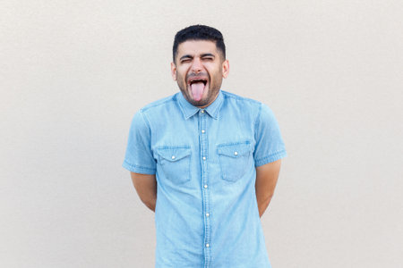 Portrait of funny crazy handsome man wearing denim shirt standing with closed eyes and showing tongue out, demonstrates childish behavior. Indoor studio shot isolated on gray background.の写真素材