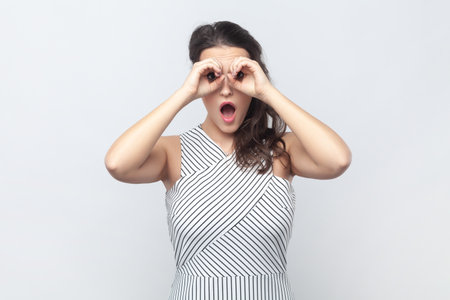 Portrait of surprised interested brunette woman making binoculars gesture with hands and looking amazed at camera, wearing striped dress. Indoor studio shot isolated on gray background.の写真素材