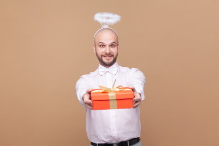 Portrait of friendly bald bearded man with nimb over head, congratulating with Valentines Day, giving present, wearing light pink shirt and bow tie. Indoor studio shot isolated on brown background.の写真素材