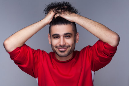 Portrait of handsome attractive smiling bearded man with curly hair, wearing red jumper, standing looking at camera, collected his hair. Indoor studio shot isolated on gray background.の写真素材