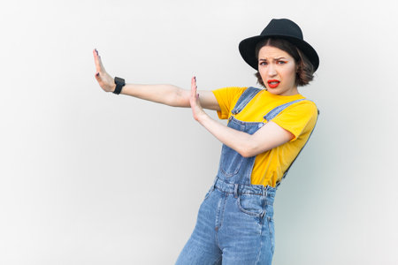 Portrait of scared shocked hipster woman in blue denim overalls, yellow T-shirt and black hat, expressing fear, showing stop gesture with both hands. Indoor studio shot isolated on gray background.の写真素材