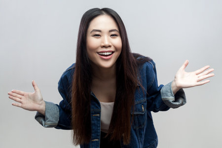 Portrait of positive brunette woman in blue denim jacket standing standing with spread hands, looking at camera and laughing. Indoor studio shot isolated on gray background.の写真素材