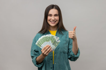 Smiling dark haired woman holding many euro banknotes and showing thumb up to camera, satisfied of winning lottery, wearing casual style jacket. Indoor studio shot isolated on gray background.の写真素材