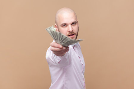 Portrait of handsome rich bald bearded man holding out dollar banknotes, looking at camera, giving money to you, wearing light pink shirt and bow tie. Indoor studio shot isolated on brown background.の写真素材