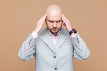 Portrait of sick ill unhealthy bald bearded man standing with hands on head, keeps eyes closed,...