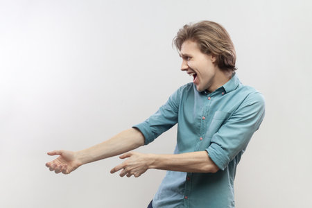 Side view portrait of strong powerful attractive young man pulling something with effort, keeps mouth open, screaming, wearing blue shirt. Indoor studio shot isolated on gray background.の写真素材