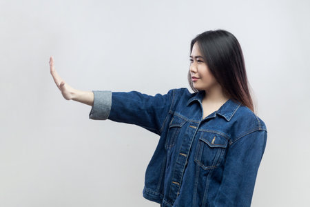 Portrait of brunette woman in denim jacket standing making stop gesture showing palm of hand, conflict prohibition warning about danger, stop bullying. Indoor studio shot isolated on gray background.の写真素材