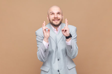 Portrait of hopeful nervous bald bearded man crossing fingers for luck, hope for better, ritual, looks up with hopeful look, wearing gray jacket. Indoor studio shot isolated on brown background.の写真素材