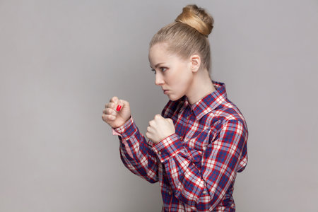 Side view portrait of angry woman with bun hairstyle standing with clenched fists, being ready to attack somebody, wearing checkered shirt. Indoor studio shot isolated on gray background.の写真素材