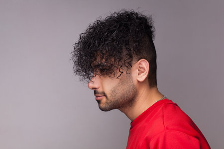Side view portrait of attractive handsome young adult bearded man with curly hair, wearing red jumper, looking ahead, showing his hairdo. Indoor studio shot isolated on gray background.の写真素材