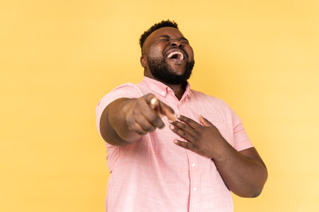Portrait of funny positive bearded young adult man wearing pink shirt laughing out loud holding belly and pointing finger on you, mockery. Indoor studio shot isolated on yellow background.の写真素材