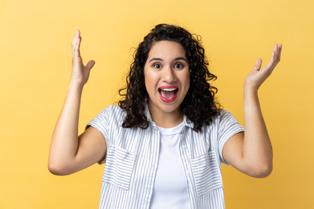 Portrait of amazed excited woman with dark wavy hair looking nat camera with excitement, keeping hands on cheeks, pleasant surprise. Indoor studio shot isolated on yellow background.の写真素材