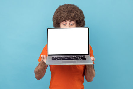 Portrait of unknown man with Afro hairstyle in orange T-shirt holding laptop in hands, hiding half of his face, copy space for promotion, looking down. Indoor studio shot isolated on blue background.の写真素材